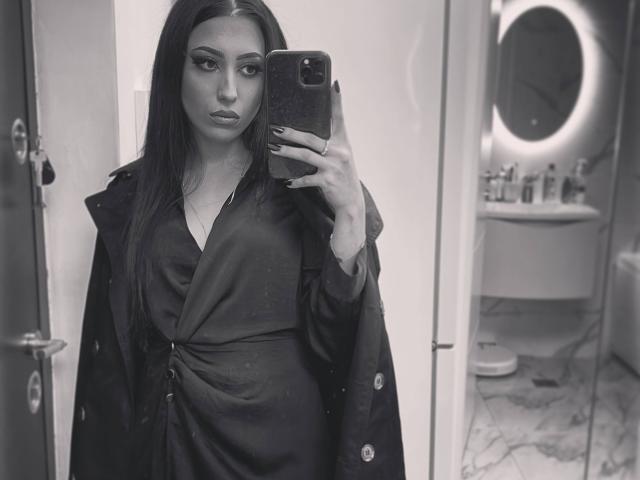 TheaOrre with long dark hair, wearing a dark dress and coat, taking a mirror selfie, showing off her pouty lips and subtle makeup, in a black-and-white photo.