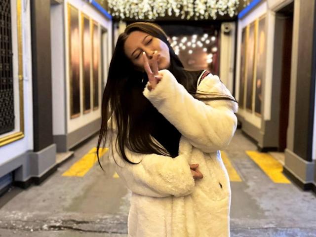 TheaOrre, with long brown hair, is in a white fluffy coat, holding up a peace sign, eyes closed, smiling softly, standing in a dimly lit hallway.