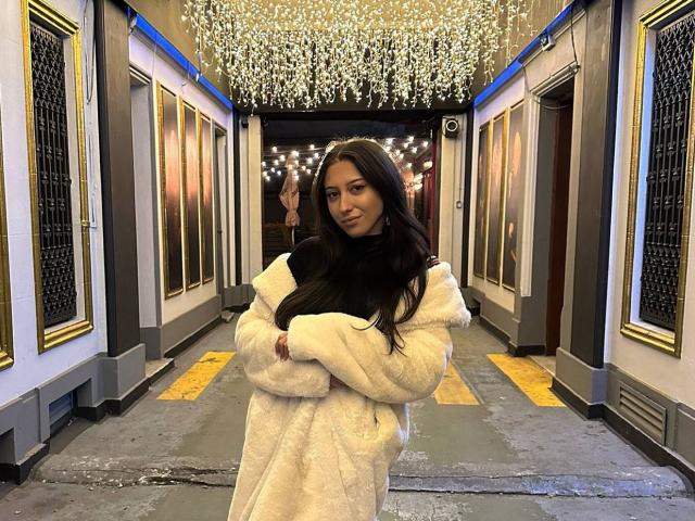 TheaOrre with long black hair, white fluffy coat, arms crossed, standing in a dark, illuminated hallway, looking at the camera, slight smile, street lights above.
