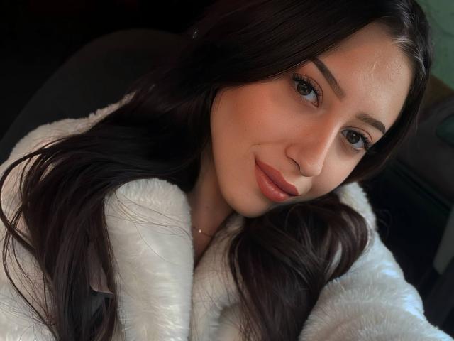 TheaOrre has long dark brown hair, brown eyes, and a slight smile. She's wearing a fluffy white coat, looking cute with her nose slightly upturned.