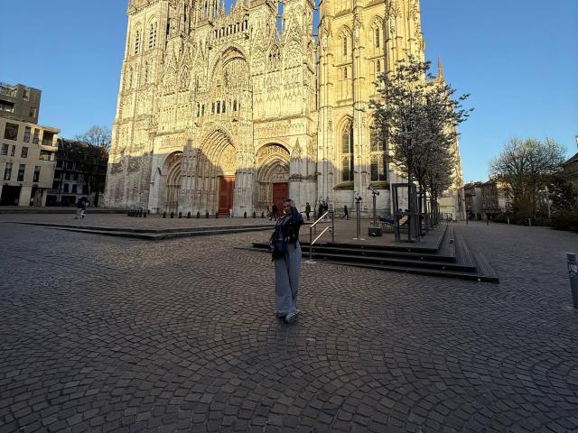TheaOrre stands in front of a huge Gothic cathedral, wearing a blue backpack and grey pants, with a small tree to the right and clear blue sky above.