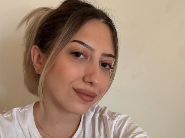 TheaOrre with light brown hair in a ponytail, wearing a white shirt, has a slight smile, brown eyes, and subtle makeup, looking slightly to the side.