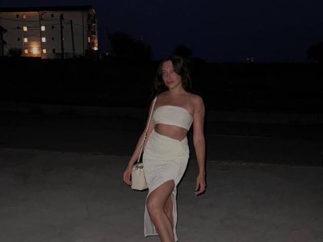 TheaOrre has tits, wearing a strapless white bandeau and a high-slit white dress, with a white handbag, standing at night with a dark blue sky and a building.