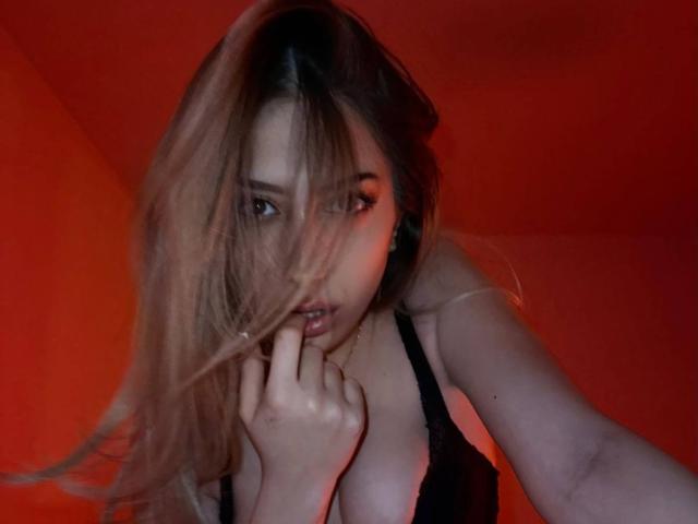TheaOrre with long brown hair, biting finger, looking seductively at camera, wearing black lingerie, red lighting, showing her tits. Her hand is near her mouth.