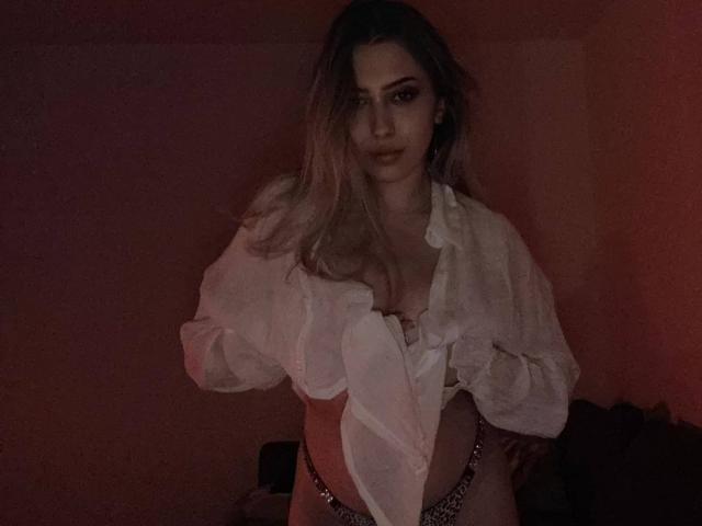 TheaOrre has long, blonde hair, wearing an open white shirt, revealing her tits, and a leopard print thong, standing in dim, red lighting, looking seductively at the camera.