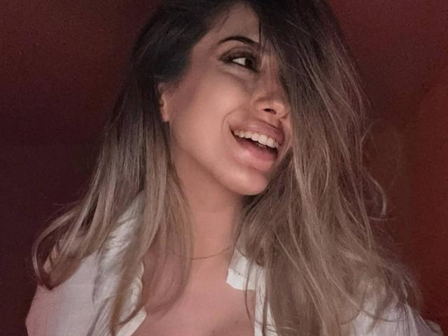 TheaOrre has long, wavy blonde hair covering one eye, smiling with her tongue slightly out, wearing a white open shirt, and a thin necklace.
