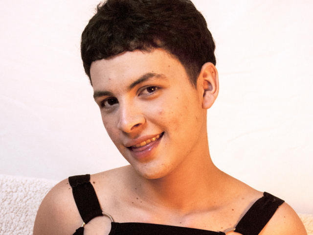 AxelLaciel has short curly black hair, brown eyes, and a slight smile with visible acne on his cheek. He wears a black strap dress against a white background.