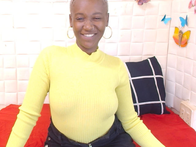 EbonyOlive69, peau foncée, souriante, portant un pull col roulé jaune texturé, un pantalon noir et des boucles d'oreilles dorées, assise sur un lit rouge, avec un oreiller noir à motifs blancs..