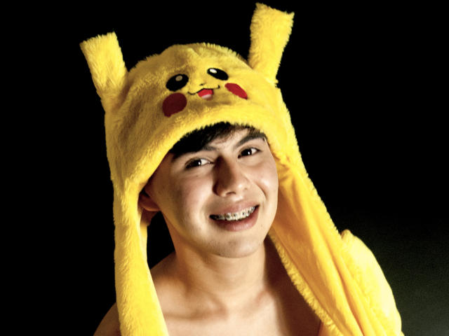 FelipeKold wearing a Pikachu yellow hat with ears and a cute face, smiling with white teeth, dark short hair, and a bare chest against a black background.