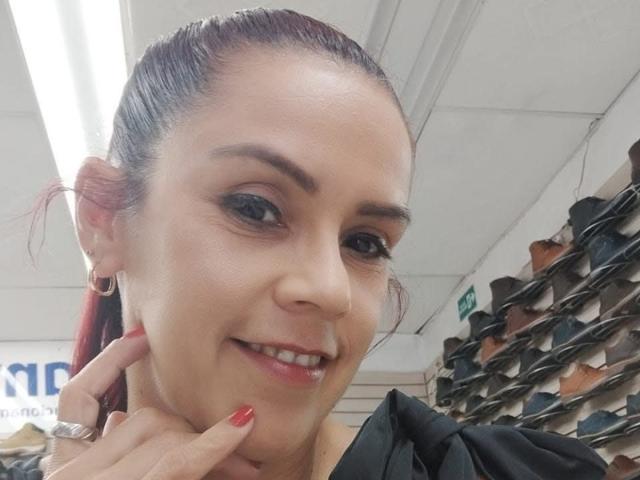 SharonBellarie with red nails, smiling, showing a cute nose ring, and ear hoops, dark eyebrows, and a slightly upped ponytail. Her hand's on her chin, in a shoe store.
