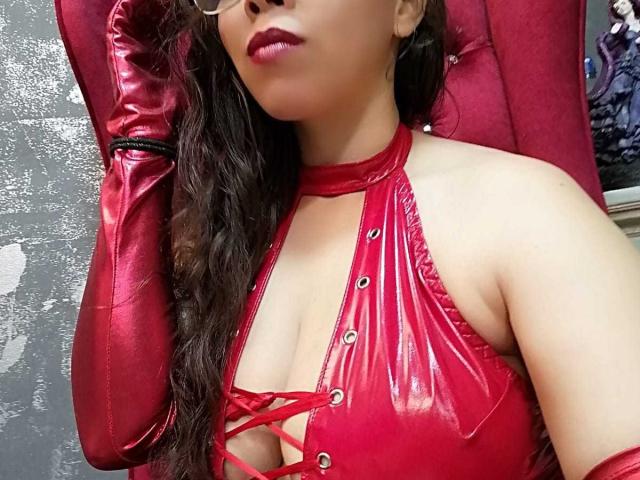 BarbaraLeythonn con lencería de látex roja brillante, corpiño con cordones y guantes a juego, mostrando sus senos, con cabello castaño ondulado y lápiz labial rojo, tocándose la cabeza..