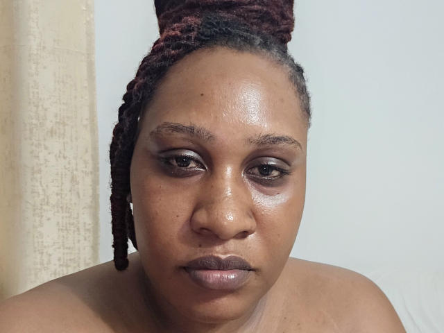 PrettyZuri has dark brown skin, glossy shiny face, and red maroon braided hair. She's topless, showing her bare shoulders, with subtle makeup and a neutral expression.