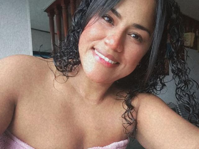 IvannaDuran, with curly black hair, smiling at the camera, has tan skin, brown eyes, and is wrapped in a pink towel, showing her bare shoulders and small tits.