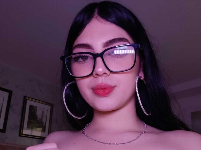 IsabellaaRossi with dark hair, black glasses, and pink lips, wearing large hoop earrings and a silver necklace, has a cute and confident expression. Her tits are not visible.