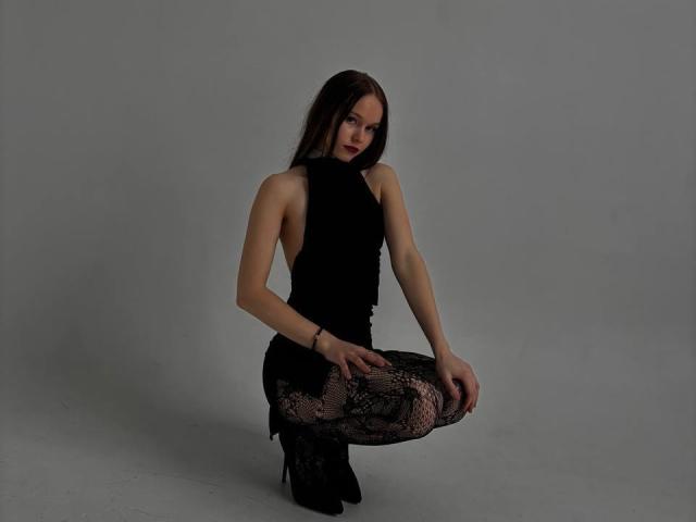 LacreshaZank has long brown hair, wears a black sleeveless top and black lace-patterned leggings, squatting with hands on knees, looking at camera with a slight smirk.