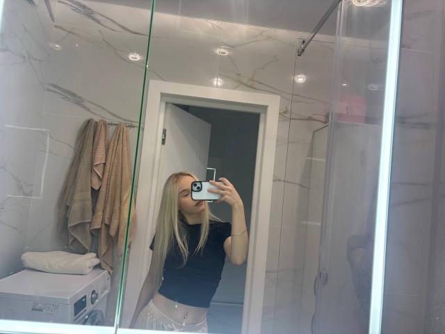 LacreshaZank with blonde hair, black top, and white pants, taking a mirror selfie, showing her ass in a bathroom with marble walls and towels hanging.