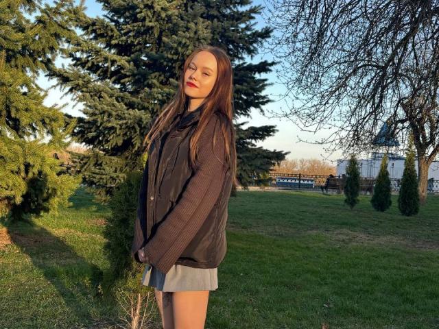 LacreshaZank with long brown hair, wearing a black sweater and gray skirt, standing in a grassy yard with a tall evergreen tree and bare branches in the background.