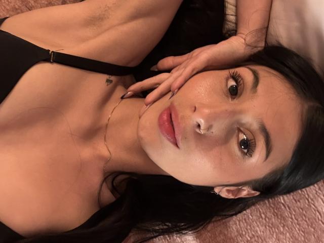 SophieBeaumont with dark hair, black top, and pink lips, lying on a pink textured surface, hand on cheek, brown eyes, and a small tattoo near her collarbone.