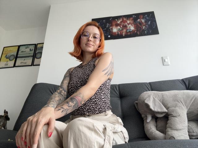SarahRousen with red hair, glasses, leopard print top, tattooed arm, sitting on gray couch, beige pants, red nails, confident smile, gray pillow, stuffed elephant.
