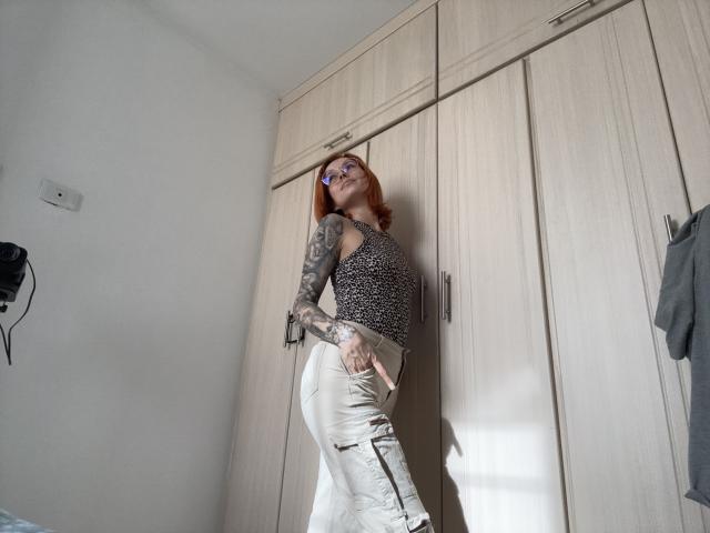 SarahRousen with red hair, tattoos on her arms, wearing glasses, a leopard print top, and white ripped jeans, standing against wooden cabinets, looking up with a slight smile.