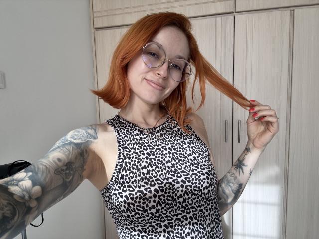 SarahRousen with red hair, glasses, and tattoos, holding a strand of hair, wearing a leopard print top, showing her tits. She's smiling and has red nails.