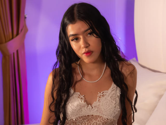 Abby Jaimes mit langem, schwarzem, gewellten Haar, trägt eine weiße Spitzen-Bralette, eine Perlenkette und roten Lippenstift, blickt leicht nach unten, in einem sanft beleuchteten Schlafzimmer. Die Brüste sind sichtbar..