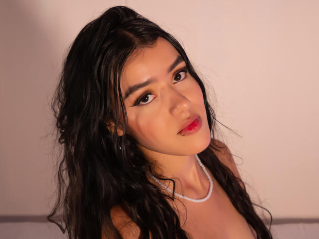 AbbyJaimes heeft golvend zwart haar, bruine ogen en rode lippenstift. Ze draagt een parelketting en kijkt iets omhoog met een subtiele glimlach. Haar borsten zijn gedeeltelijk zichtbaar..