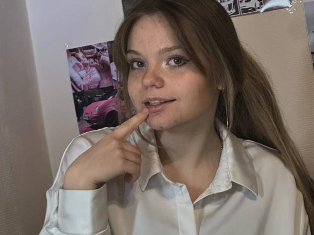 DeliaRodrick with long brown hair, wearing a white button-up shirt, has a finger to her slightly parted lips, showing a small amount of drool on her chin.