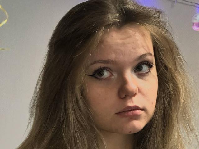 DeliaRodrick has light brown, wavy hair, a nose piercing, and is looking to the side with winged eyeliner, a slightly pouty expression, and a small mole under her lip.