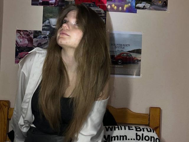 DeliaRodrick with long brown hair, white jacket, black top, and a slightly annoyed expression, sitting on a wooden bed, her tits partially covered, in a dimly lit room.