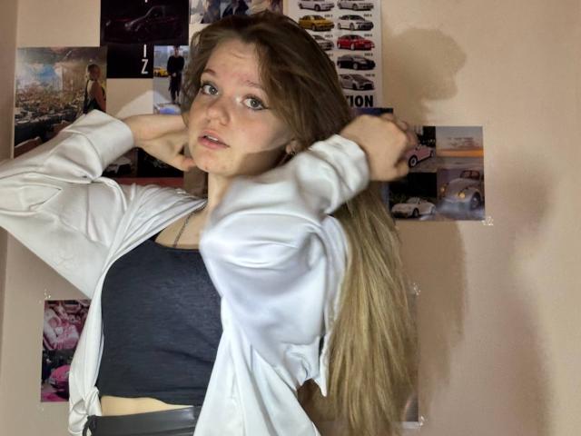 DeliaRodrick with long blonde hair, blue eyes, and a slightly pouty face, wearing a white jacket over a dark sports bra, hands in hair, standing with a curious expression.