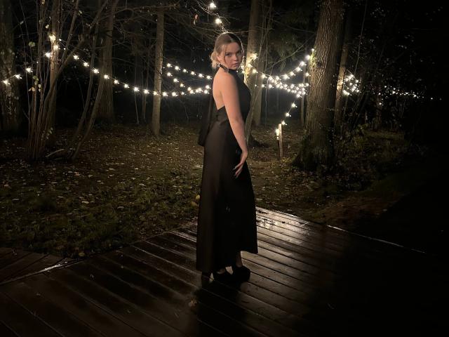 DeliaRodrick stands on a wooden deck at night, wearing a black, sleeveless, floor-length dress, showing her bare ass, with string lights and trees in the background.