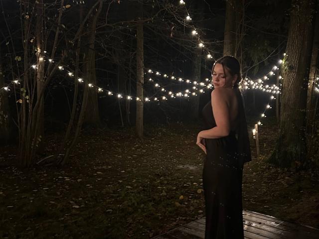 DeliaRodrick stands in a dark forest wearing a black strapless dress, looking over her shoulder with her hands on her pregnant belly, surrounded by string lights.