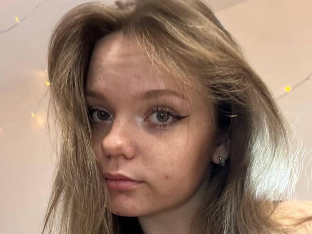 DeliaRodrick has long, dirty blonde hair, green eyes with black eyeliner, and a nose piercing. She's looking slightly to the side with a neutral expression.