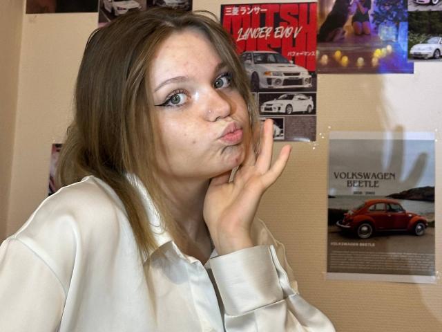 DeliaRodrick, puffy lips, making a pouty face, hand on cheek, brown hair, white shirt, freckles, subtle makeup, car posters in background, casual selfie.