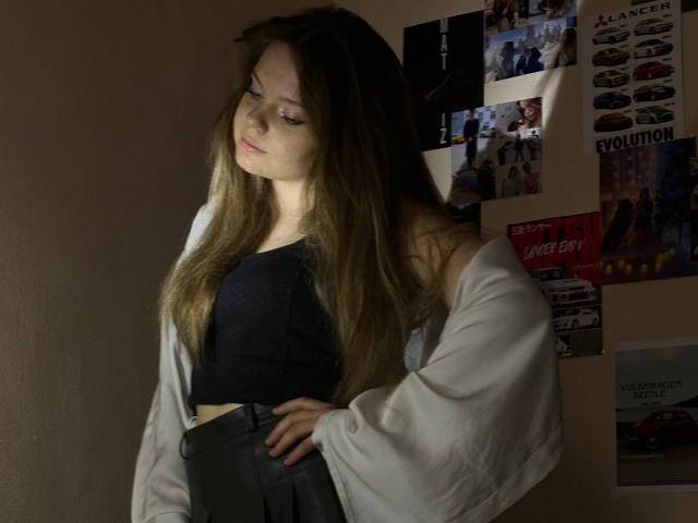 DeliaRodrick with long brown hair, off-shoulder white shirt, black crop top, and gray pants, standing in dim light, hands on hips, looking down, soft shadow on face.