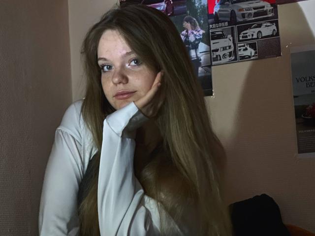 DeliaRodrick with long brown hair, blue eyes, and freckles, wearing a white shirt, resting her head on her hand, looking slightly to the side with a serious expression.