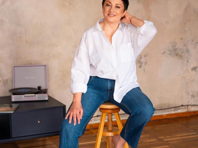 EmilyLlove is sitting on a wooden stool, wearing a white shirt with a pocket and blue jeans, smiling with one hand in her hair. Her ass is cute.