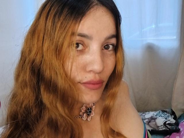 ValeriVega has long, wavy, brown-to-blonde ombre hair, brown eyes, pink lipstick, and a pink flower necklace, with freckles on her face, looking at the camera.