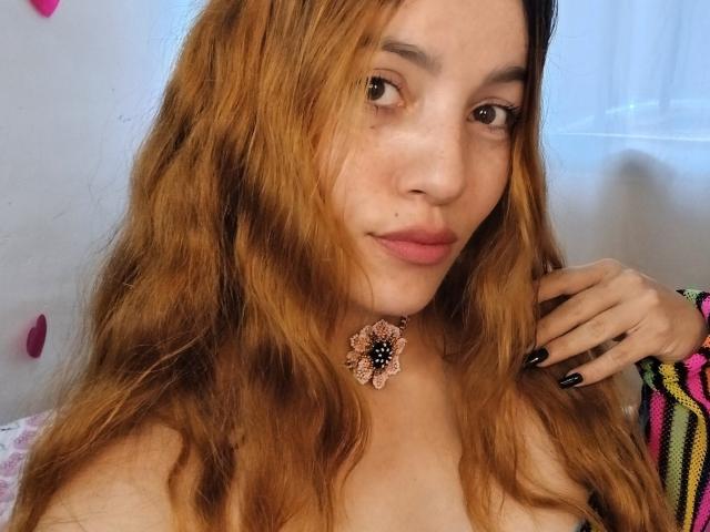 ValeriVega, com cabelo castanho ondulado, colar de flores rosa, unhas pretas e lábios rosa, olhando para a câmera, mostrando seios pequenos. Luz suave, ambiente casual..