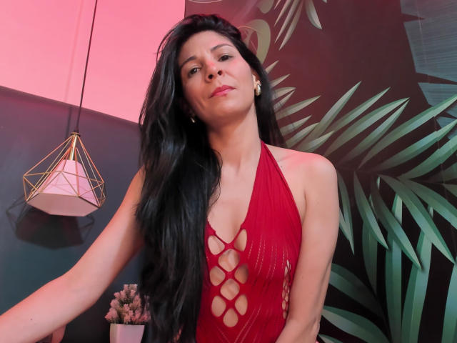 EliseBell, con lunghi capelli neri, indossa un abito rosso a fascia con motivi traforati che mostrano il seno, in piedi di fronte a un muro illuminato di rosa, con uno sfondo di foglie verdi e una lampada geometrica sospesa..