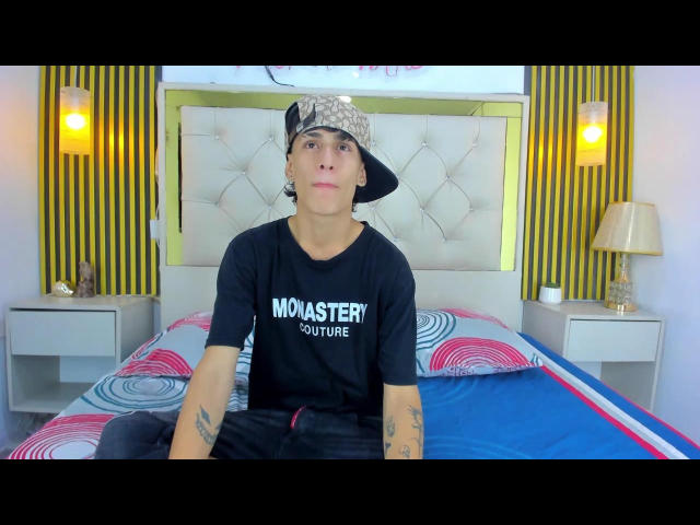 JustXJake, con tatuajes, vistiendo una camiseta negra que dice "MOUSTERUTURE" y un gorro puesto al revés, sentado en una cama con sábanas de colores y patrones circulares, mirando a la cámara..