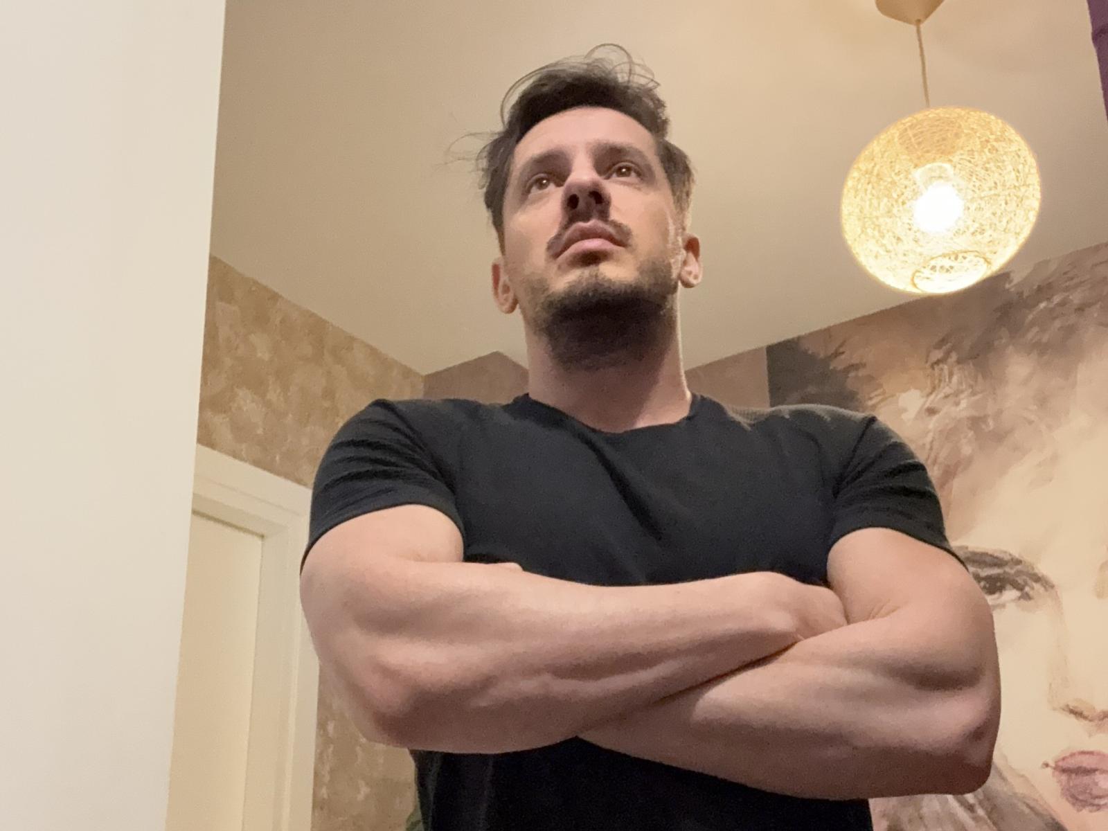 O Sr. Fitu, com os braços cruzados, vestindo uma camisa preta, olhando para cima, tem uma constituição física musculosa, cabelo curto e escuro, e uma barba discreta, sob uma lâmpada pendurada..