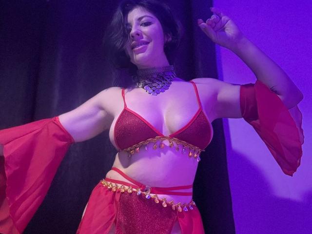 AnastasiaEvansX con un sujetador y tanga rojos brillantes, un cinturón de cadena dorada, mangas rojas, un collar negro y cabello oscuro ondulado, sonriendo bajo una luz púrpura, mostrando sus pechos..