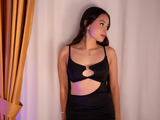 AmberJara with long dark hair, wearing a black cutout dress, showing her tits, standing against a white wall, gold curtain on the left, looking to the right.