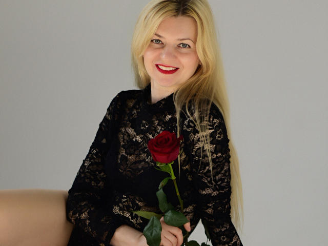 MelissaTyler with long blonde hair, wearing a black lace dress, holding a red rose, smiling with red lipstick, sitting against a plain grey background. Her tits are visible.