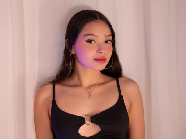 Amber Jara, avec ses longs cheveux noirs, porte un haut noir découpé, des boucles d'oreilles dorées et un collier. Son visage est illuminé d'une lumière rosée-violette, mettant en valeur sa poitrine et son joli visage..