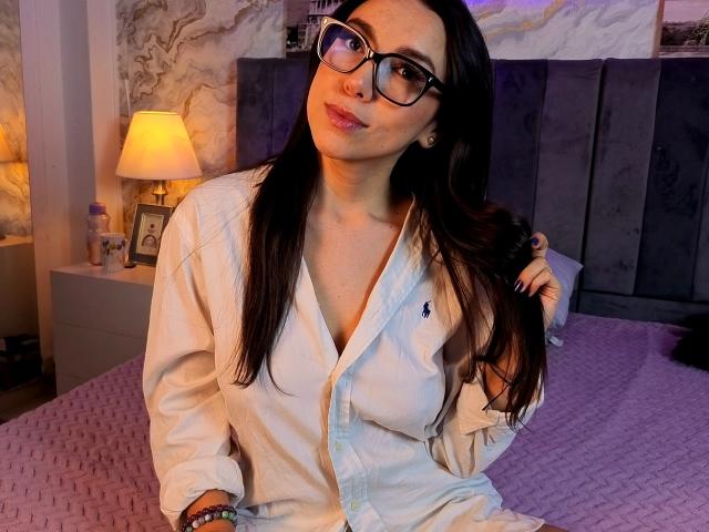 MiaReed with long black hair, glasses, and a white button-up shirt, sitting on a bed, holding her hair, showing a cute smile and small tits, in a cozy room.