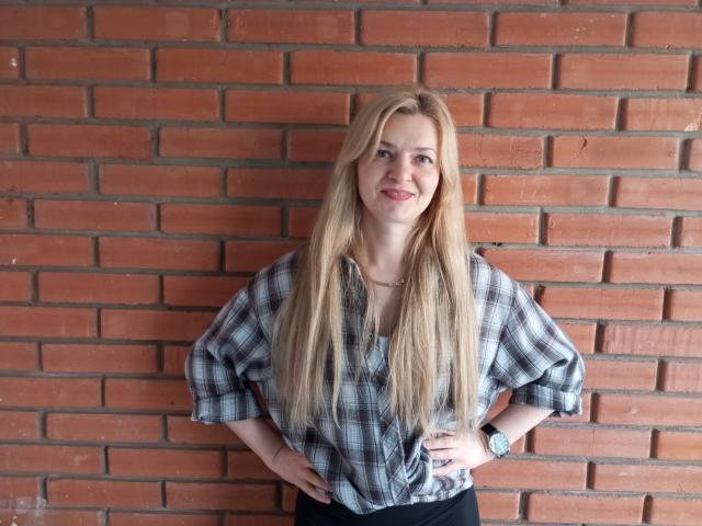MelissaTyler has long blonde hair, wearing a plaid shirt, hands on hips, smiling, with a watch on her left wrist, standing against a brick wall.