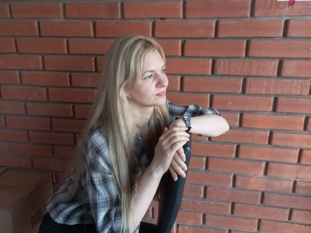 MelissaTyler with long blonde hair, plaid shirt, black chair, arms folded, wristwatch, looking to the side, brick wall in background, cardboard box on left.